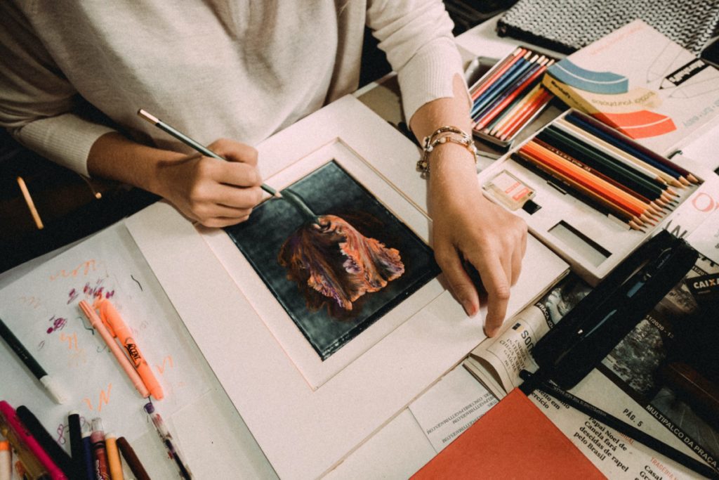 artist painting piece with an assortment of supplies on the desk around them